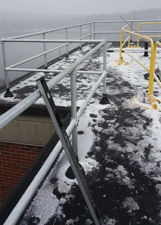 A black spirit level rests against a silver metal guardrail on a snowy, flat rooftop with a yellow rail in the distance.
