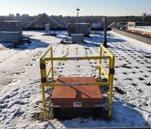 A yellow safety railing surrounds a metal roof hatch on a snowy, flat rooftop with industrial equipment in the distance.