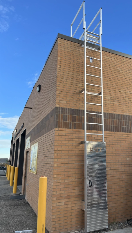 An exterior metal ladder attached to a tan brick building, featuring a security enclosure at the base and a top railing.