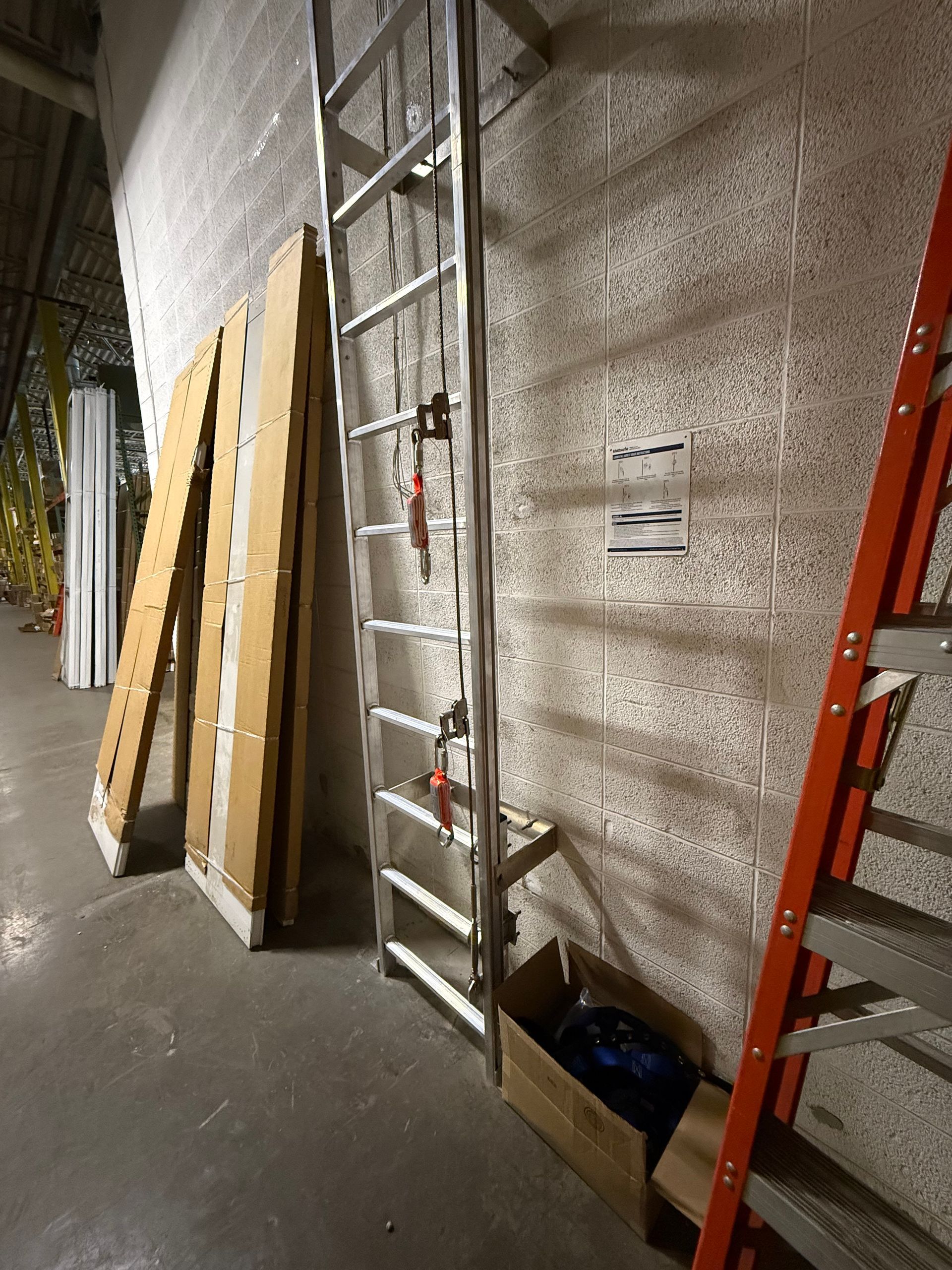 A metal ladder leans against a rough white wall, beside cardboard planks and an orange step ladder in a storage area.