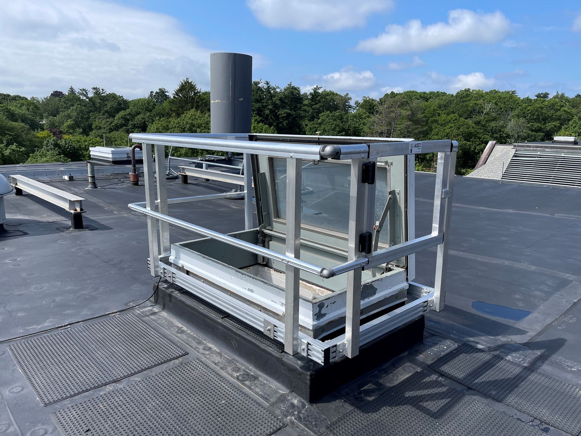 A rooftop metal safety railing surrounding a square skylight access hatch against a blue sky with scattered clouds.