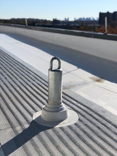 A metal roof safety anchor post installed on a gray flat rooftop under a clear blue sky.