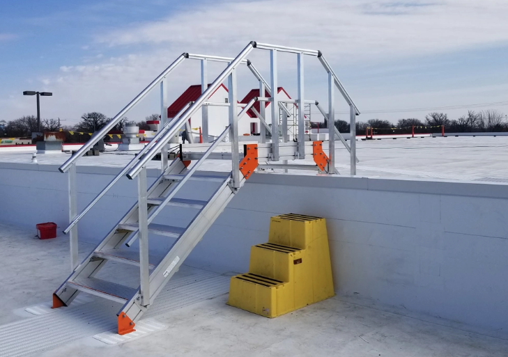 A metal crossover ladder with handrails atop a flat roof, positioned next to a small, three-step yellow plastic stool.