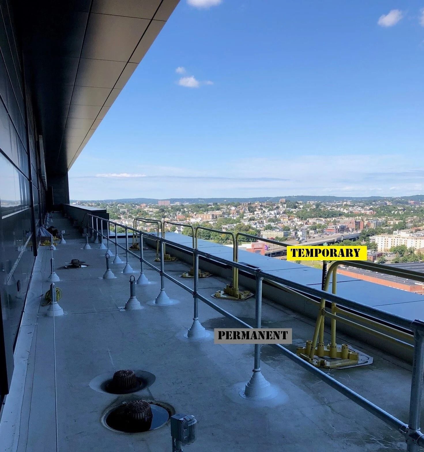 A rooftop balcony with permanent railings and a temporary yellow barrier overlooking a city skyline under a blue sky.