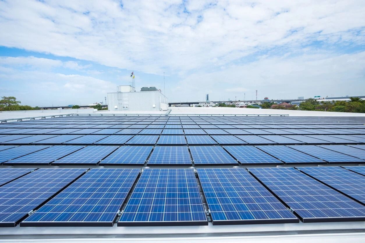 A vast array of blue solar panels installed across a flat rooftop under a bright, cloudy sky.