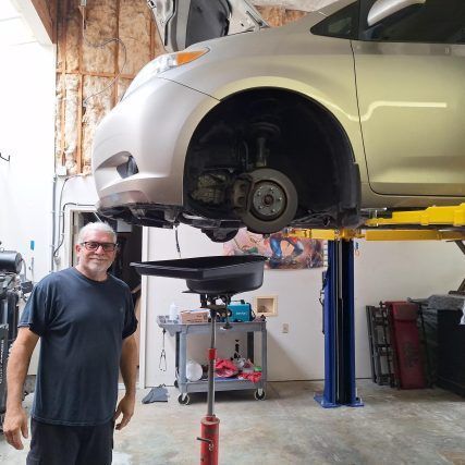 Man in a shop stands before a car on a lift; oil drain pan below.