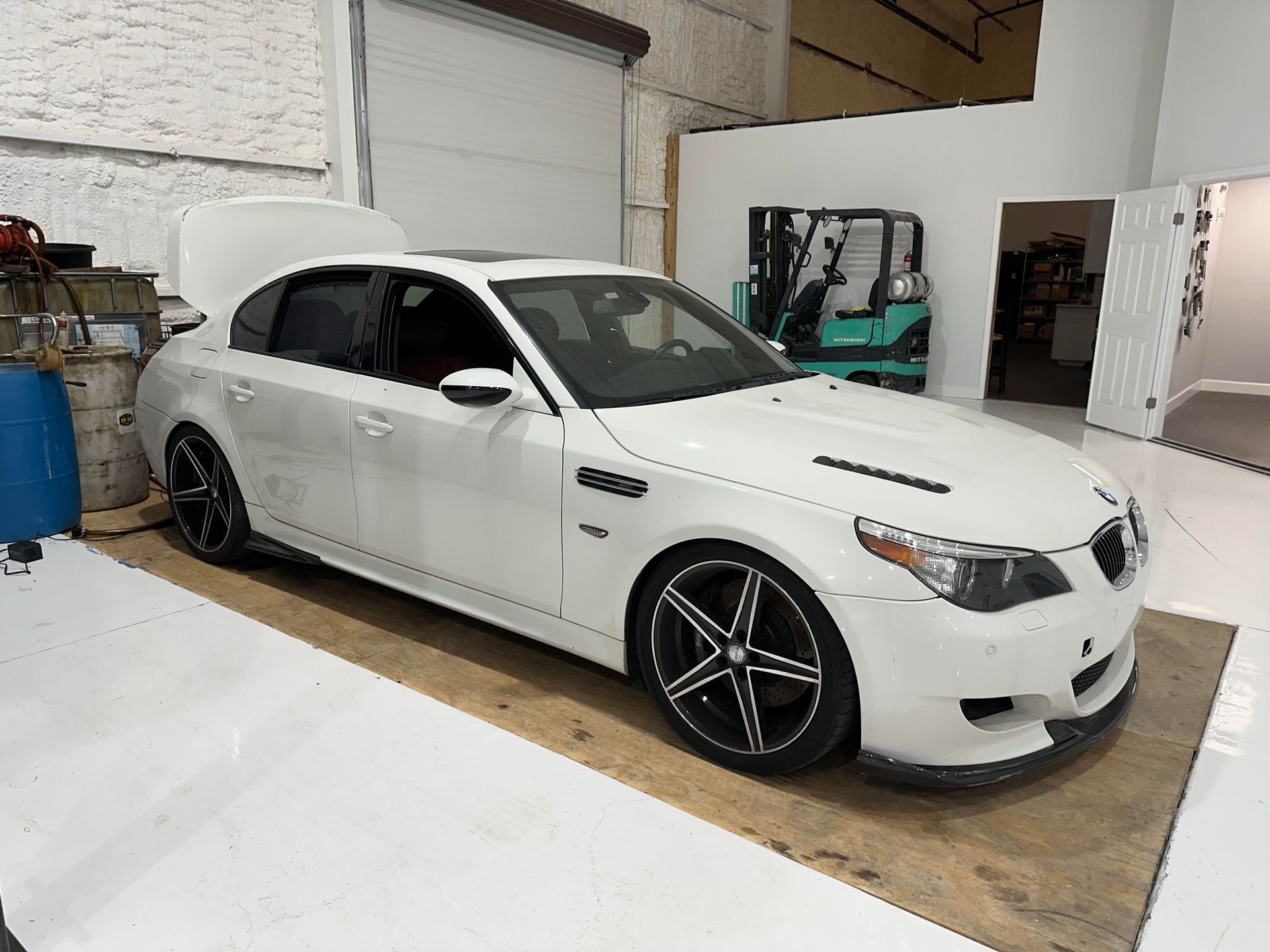 White BMW M5 sedan with black wheels, hood open, in a garage.
