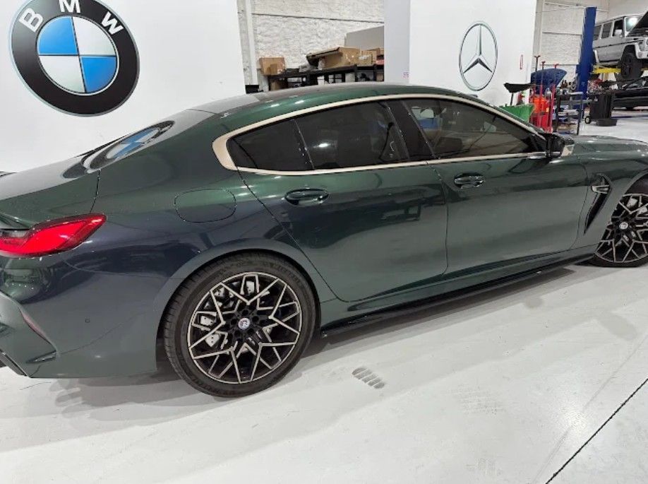 Green BMW M8 Gran Coupe in a dealership, with a BMW logo on the wall.