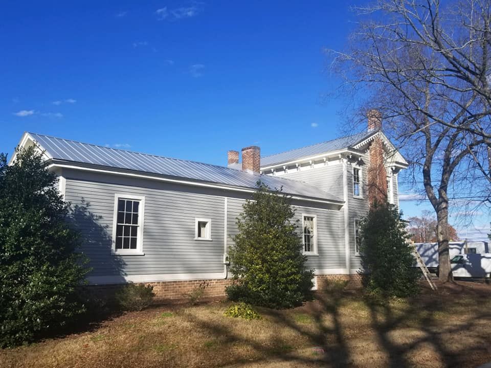 A large white house with a metal roof is surrounded by trees on a sunny day