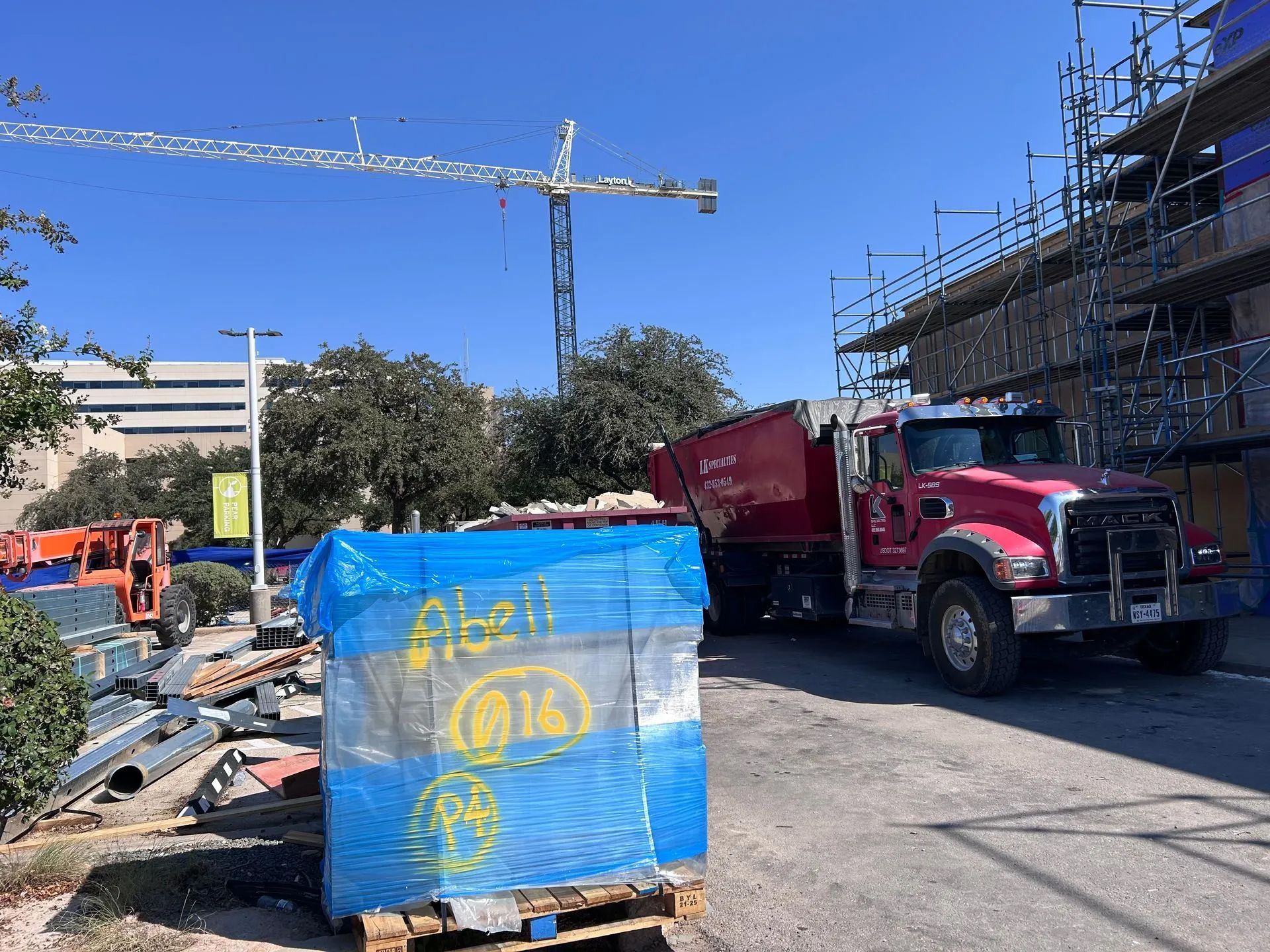 Construction site with red dump truck next to a building under construction, a crane, and materials wrapped in blue.