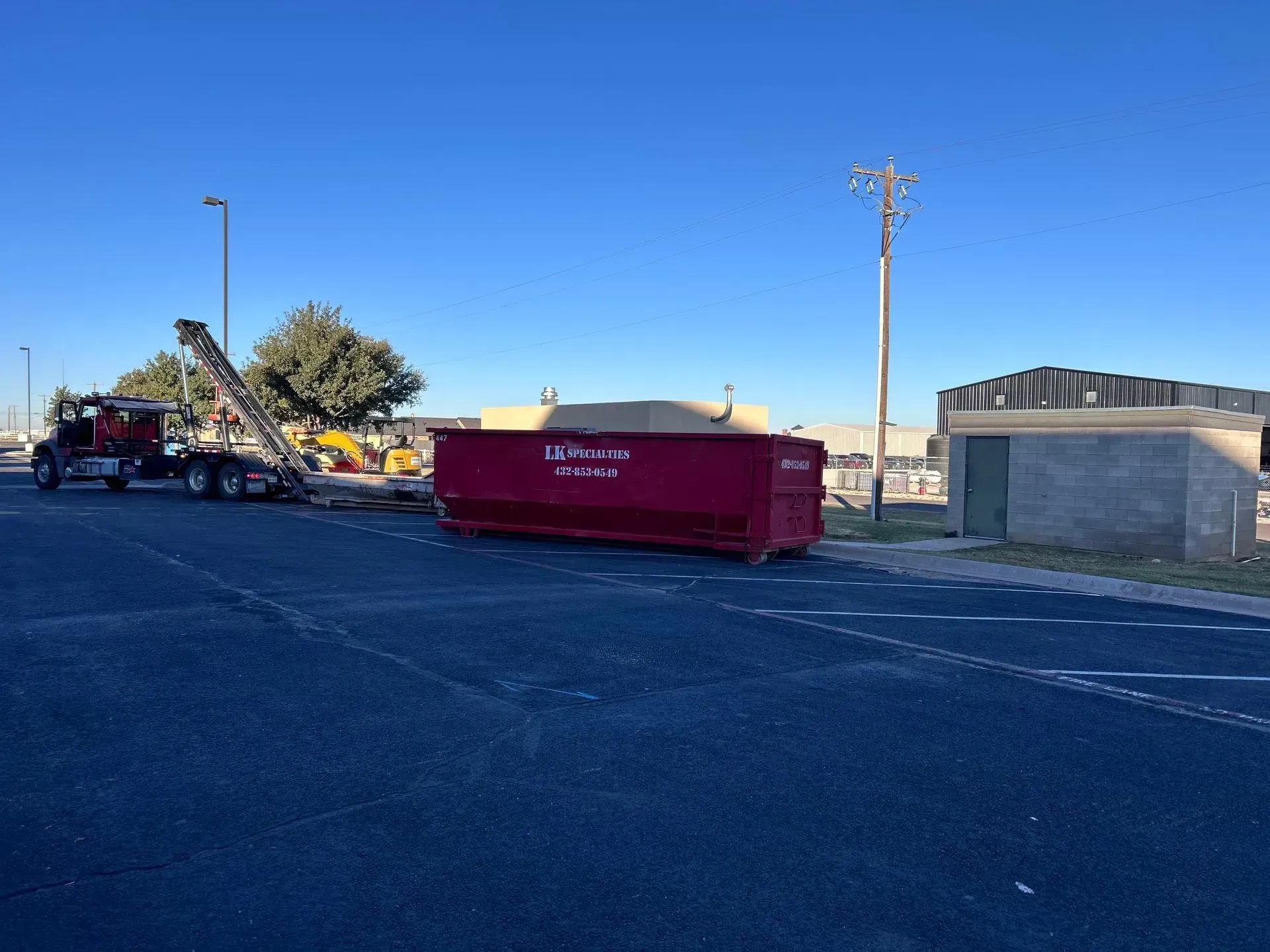 Red dumpster on asphalt next to a truck. Power lines and a building are in the background.