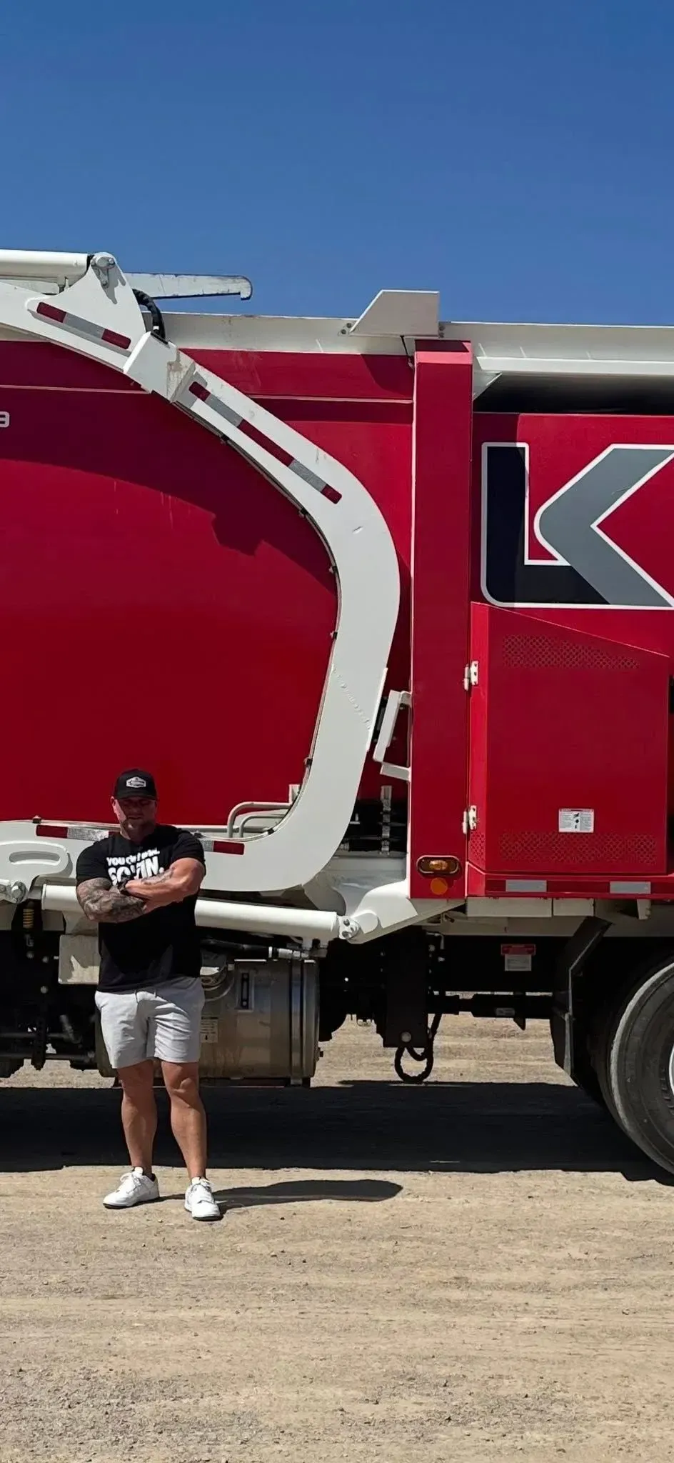 Man in shorts and t-shirt stands next to a red truck with a white arm. Blue sky and gravel.