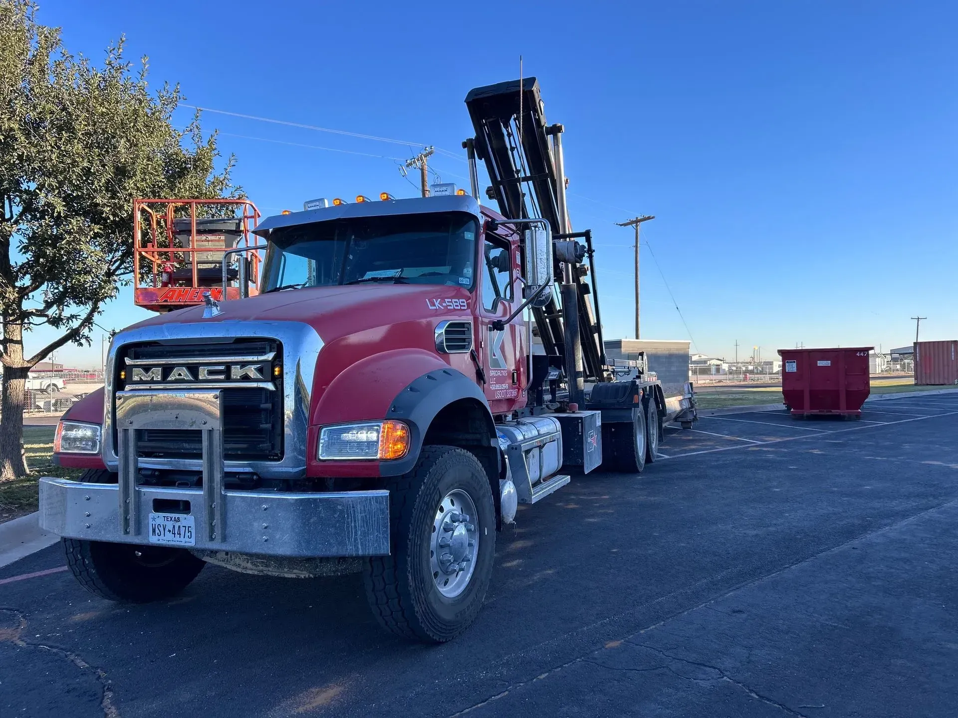 Red Mack truck with lift in a parking lot. Dumpster in the background. Sunny day.
