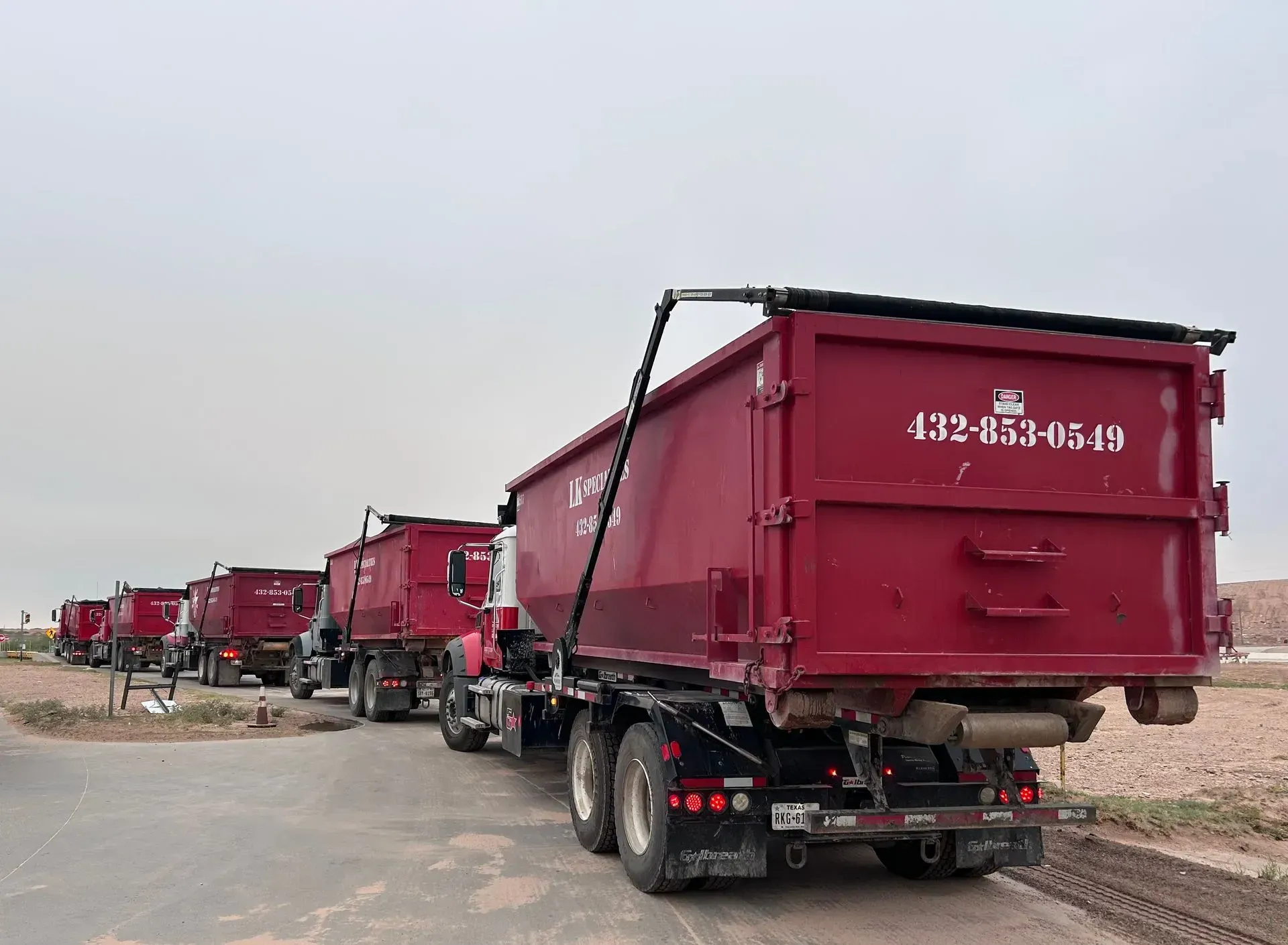 Row of maroon dump trucks on a road, under an overcast sky.