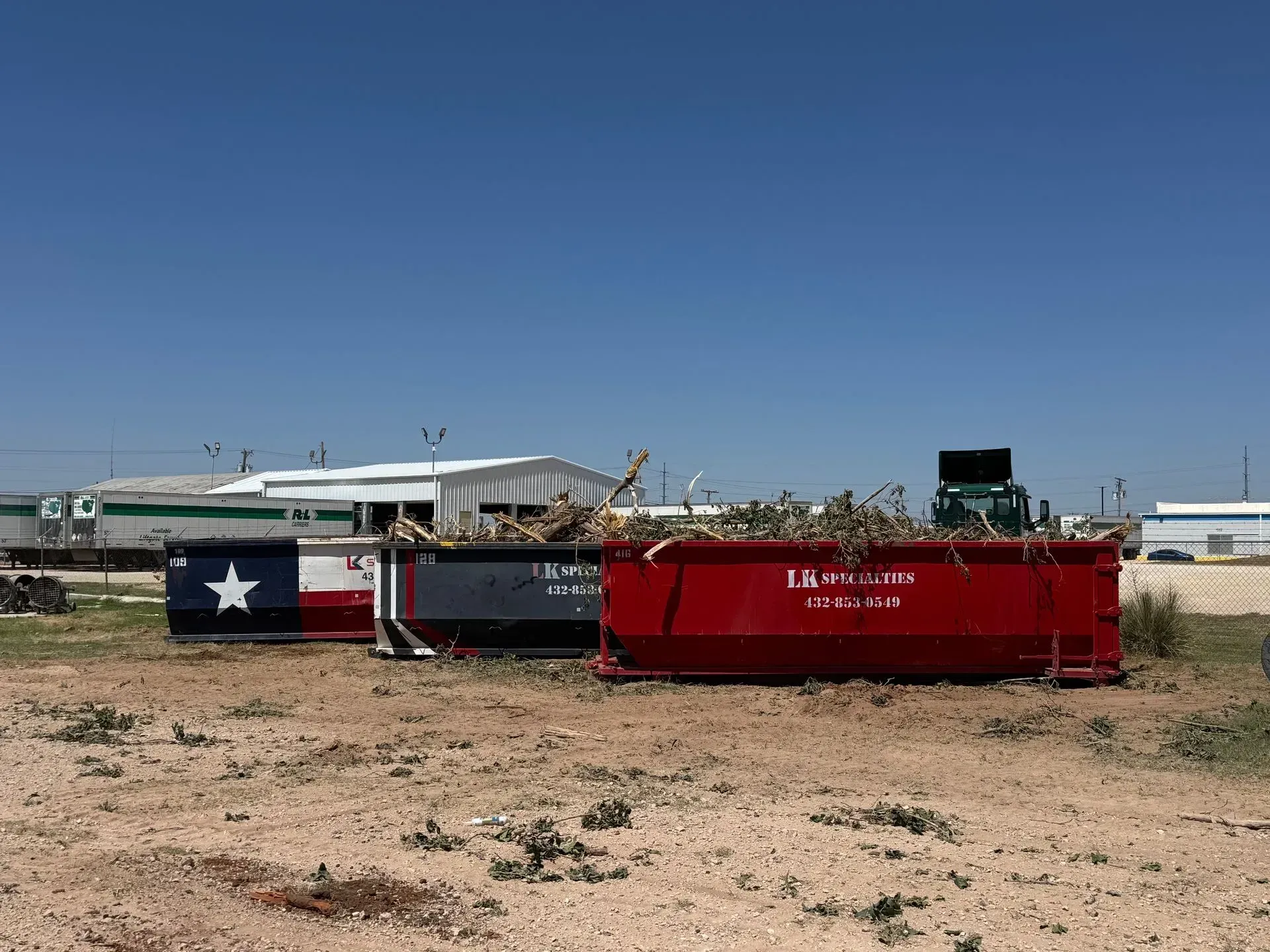 Three dumpsters filled with debris sit outdoors under a blue sky, one painted with a Texas flag.