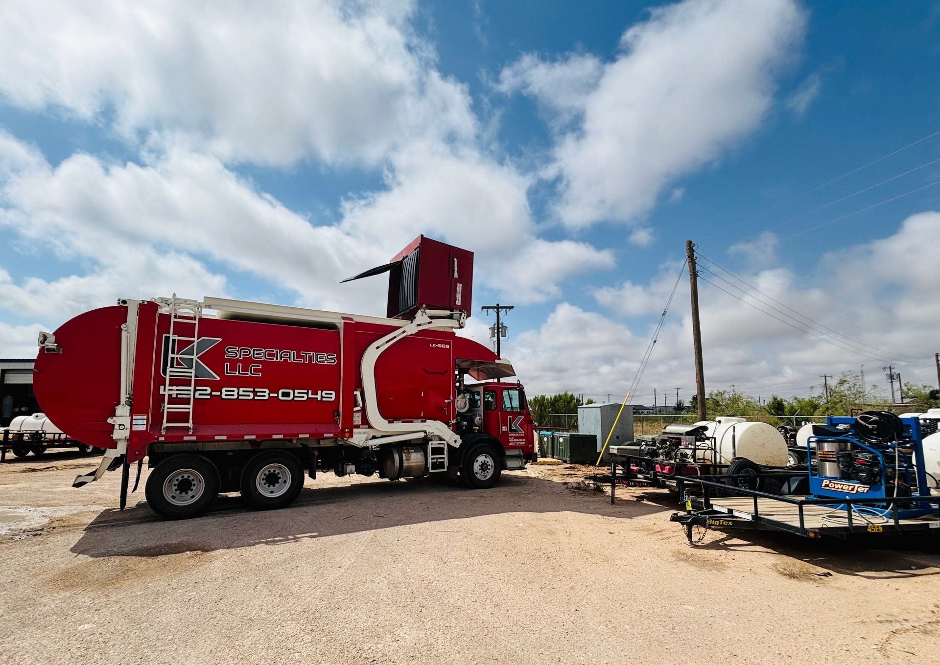 Red garbage truck parked outdoors on gravel with blue sky.