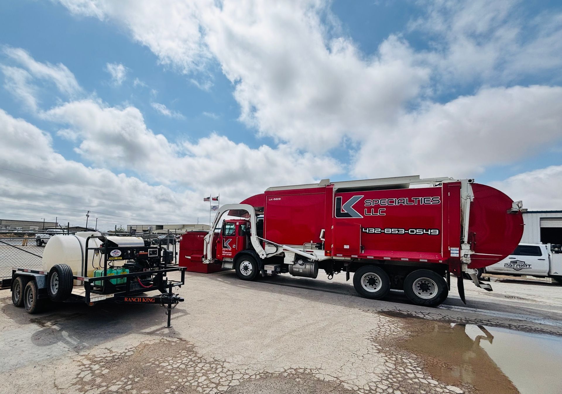 Red sanitation truck with attached trailer parked on a gravel lot under a cloudy sky.