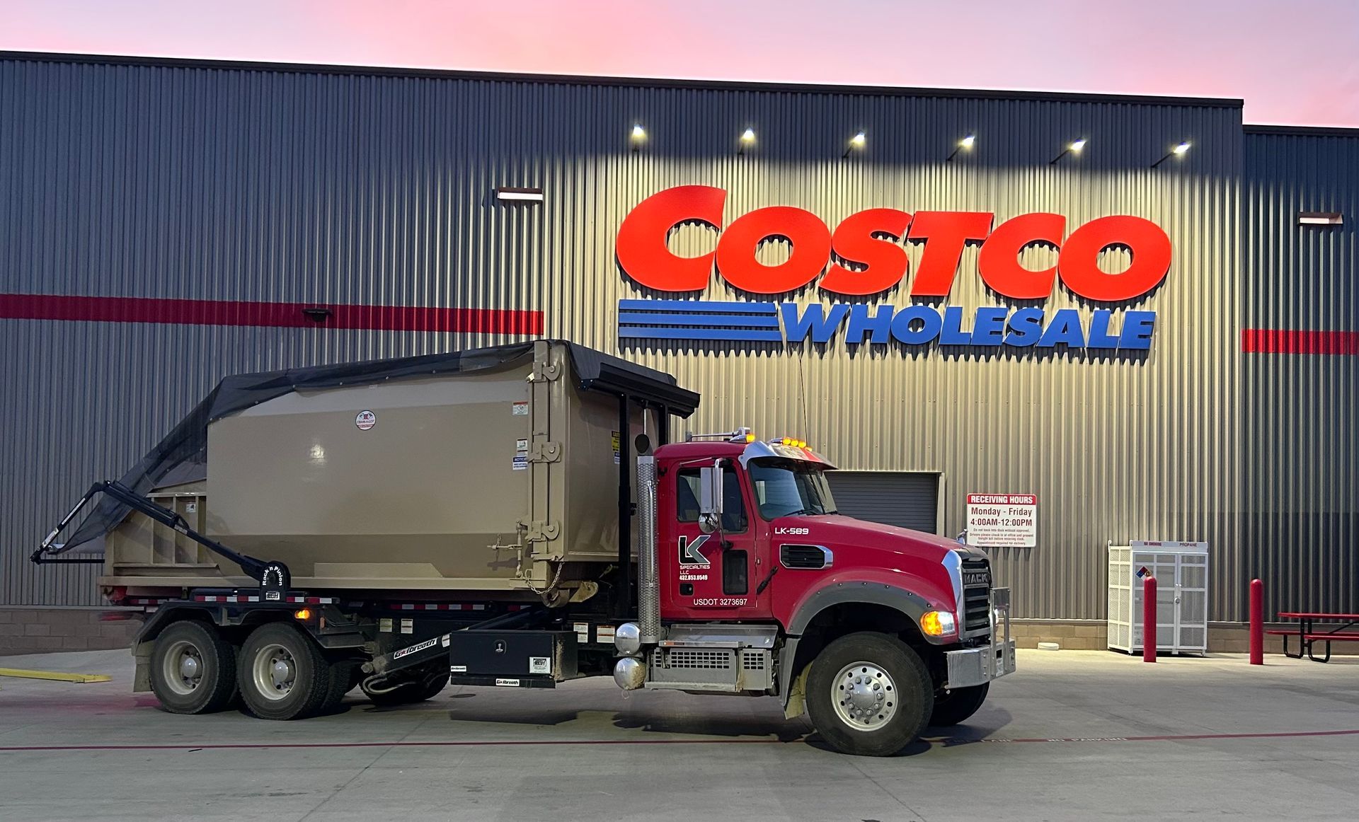 Dump truck parked in front of a Costco store. Red truck, tan bed, dusk setting, lit Costco sign.