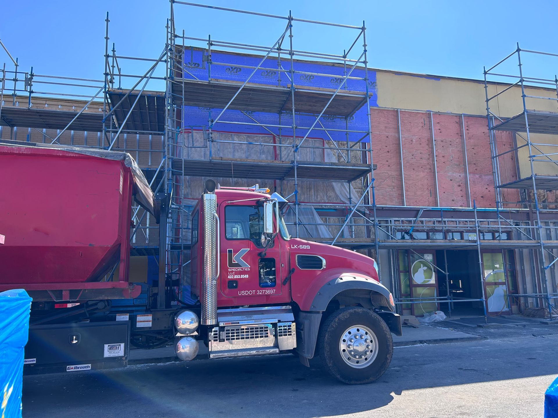 Red dump truck parked near a building under construction, scaffolding and insulation visible.