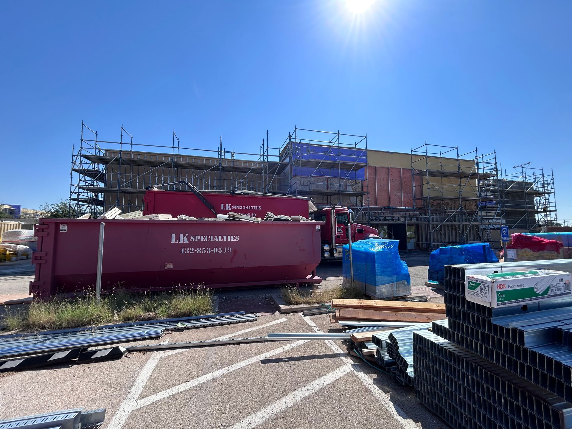 Construction site with scaffolding, dumpsters, and building materials under a sunny sky.