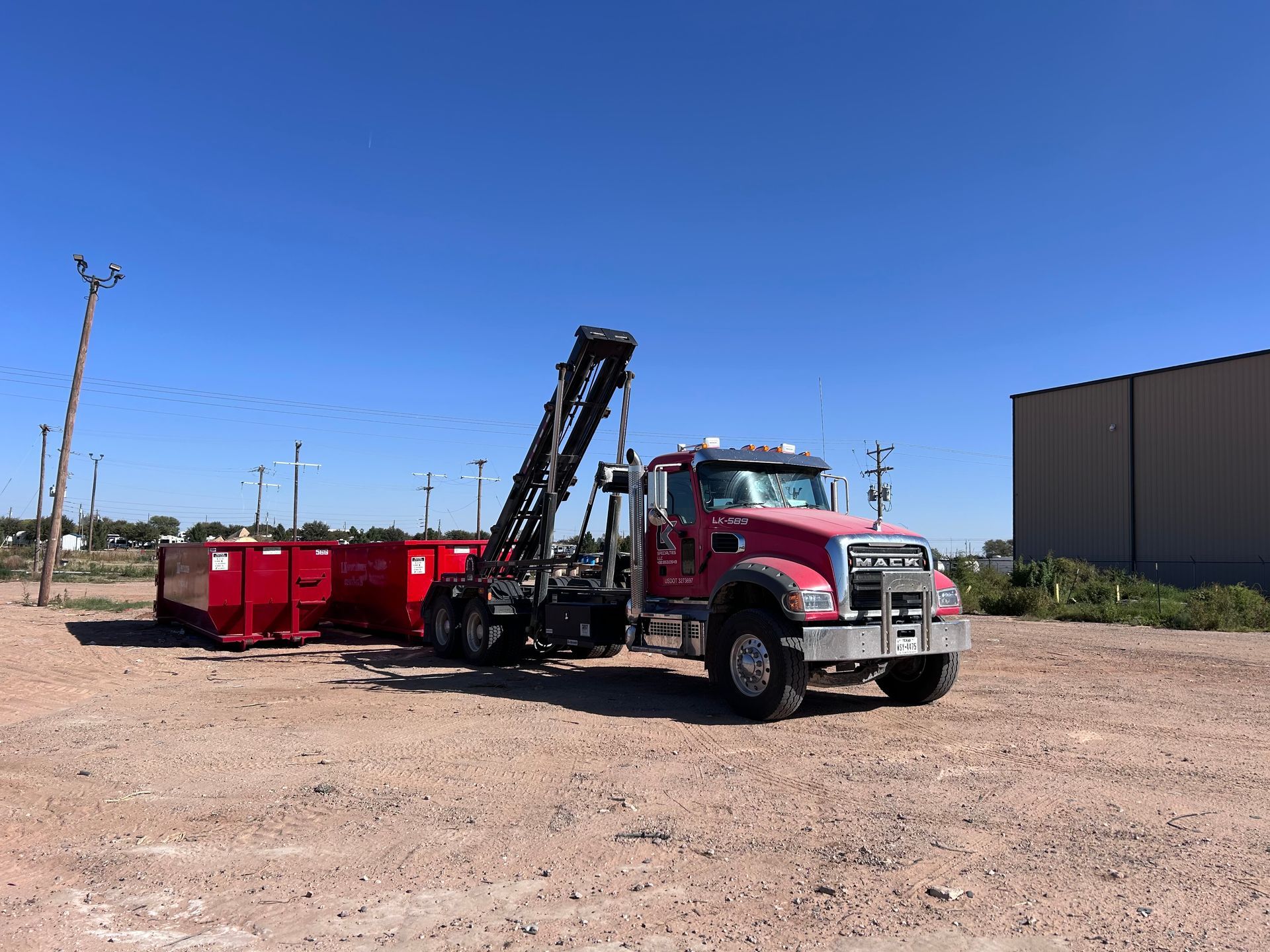 Red roll-off truck with bins on a gravel lot under a blue sky.