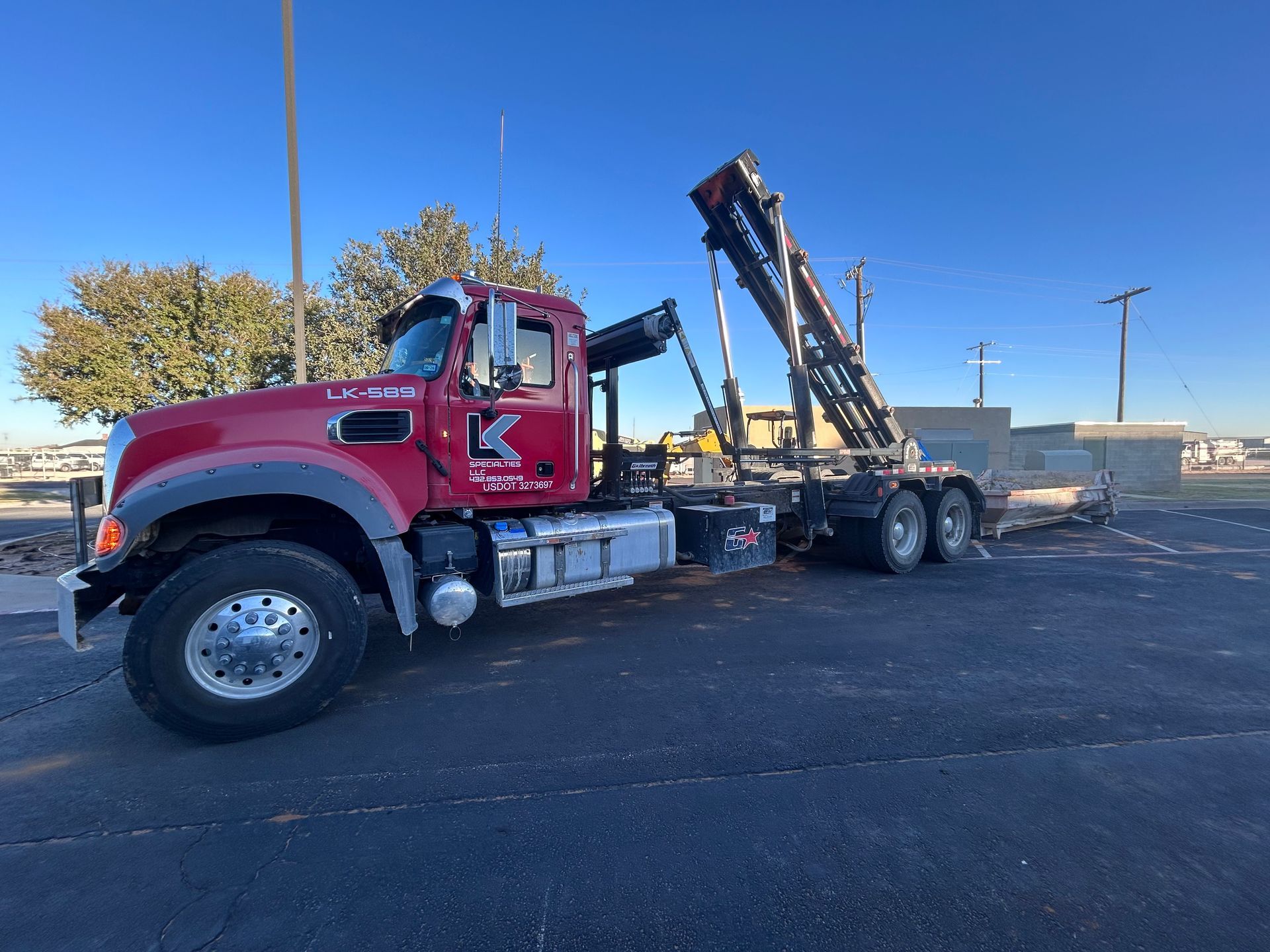 Red roll-off truck with crane lifting a container on a sunny day.