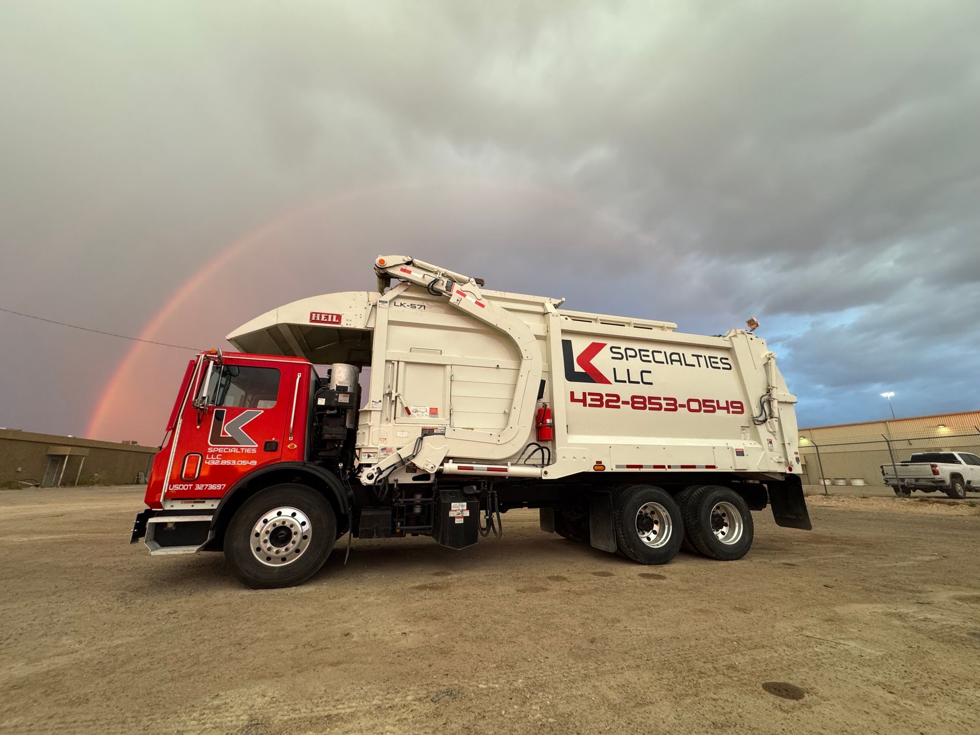 Garbage truck, red and white, parked with a rainbow in the sky.