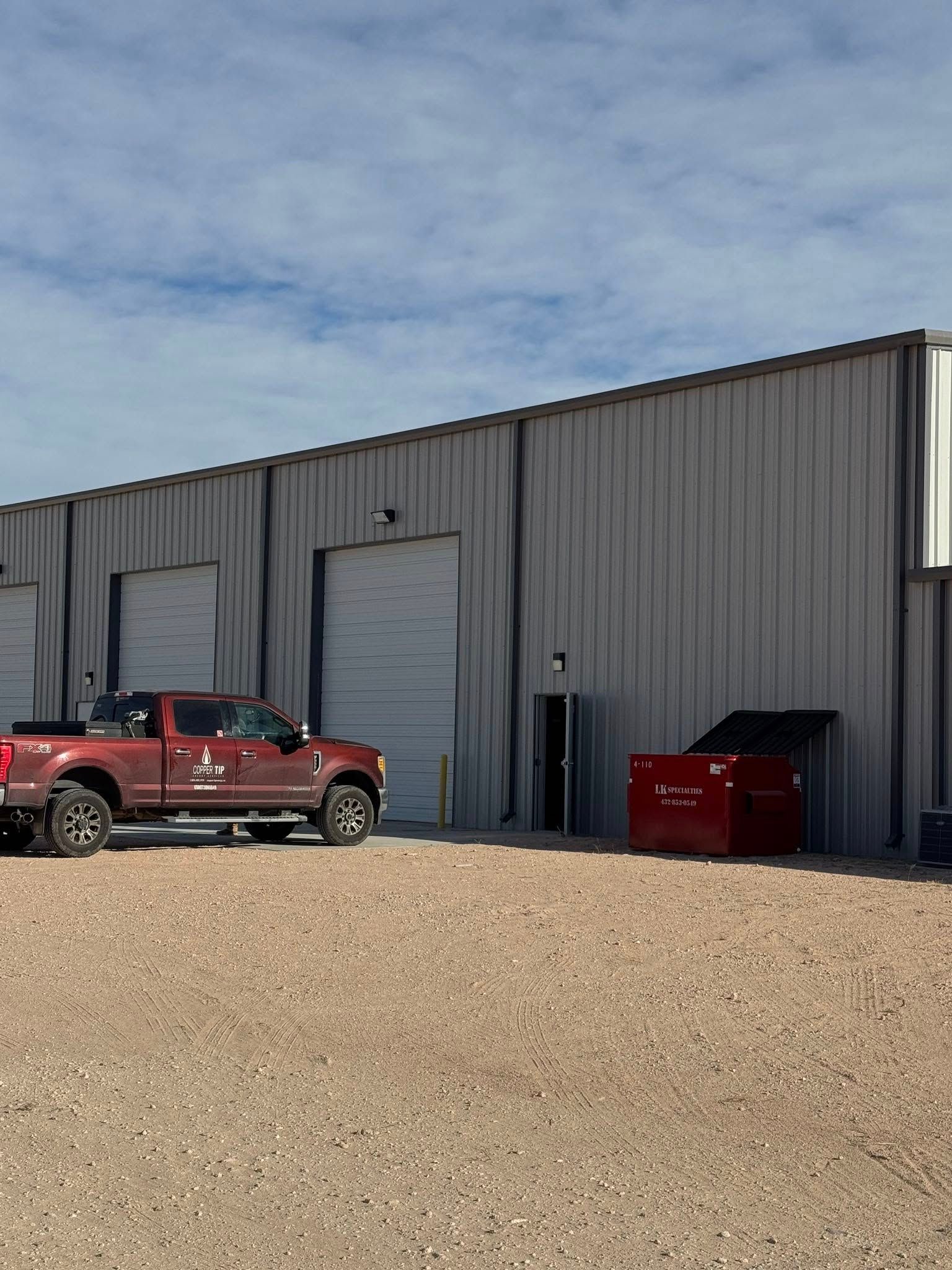 Red truck parked outside a metal building with garage doors. Ground is gravel.