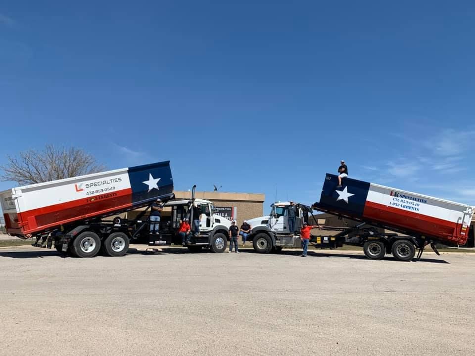 Two dump trucks with Texas flag designs on the beds, parked outdoors with people nearby.