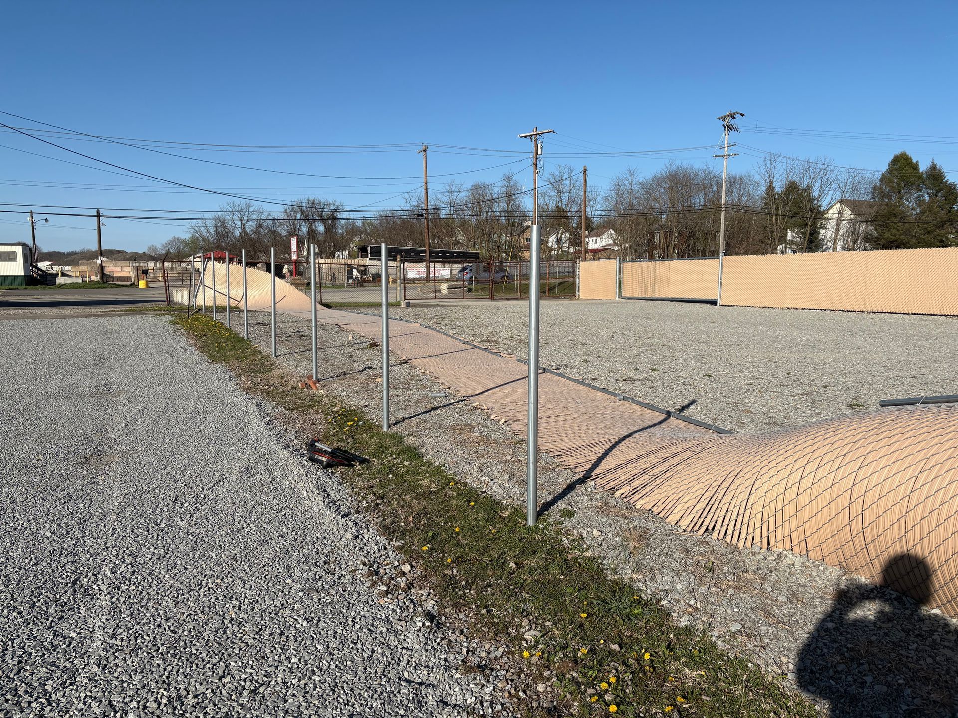 Gravel lot with a chain-link fence, tan wall, utility poles, and a clear blue sky.