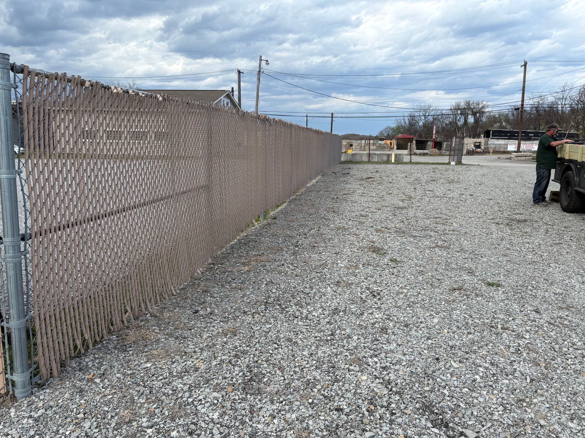 Gravel lot beside a chain-link fence under a cloudy sky, with utility poles and parked equipment.