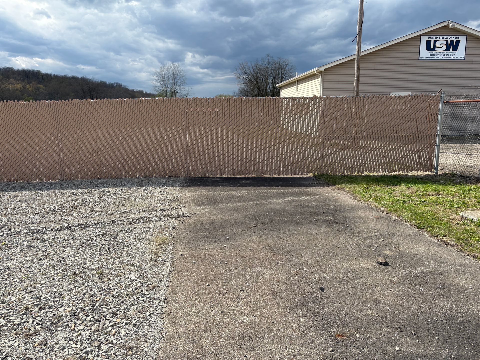 Large brown wooden fence blocking a gravel driveway beside a white building under a cloudy sky