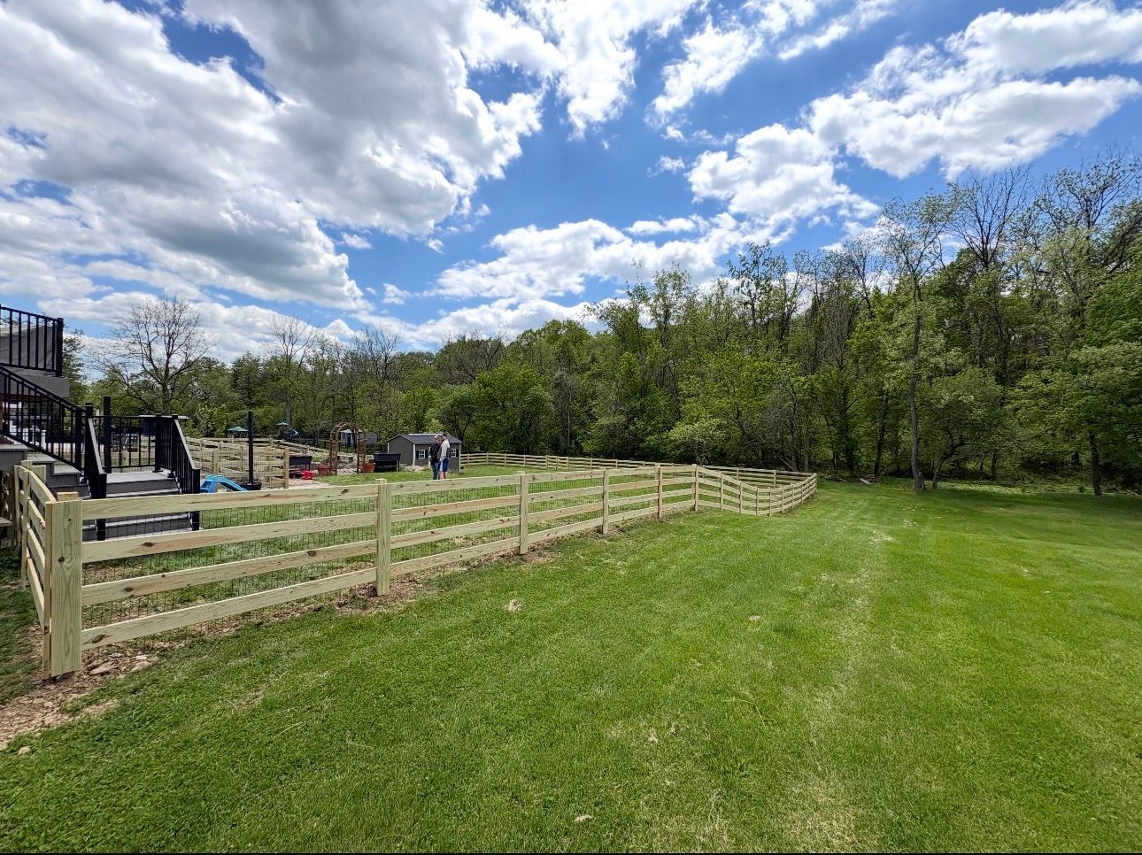 Grassy park field beside a wooden fence, trees, and a playground under a cloudy blue sky