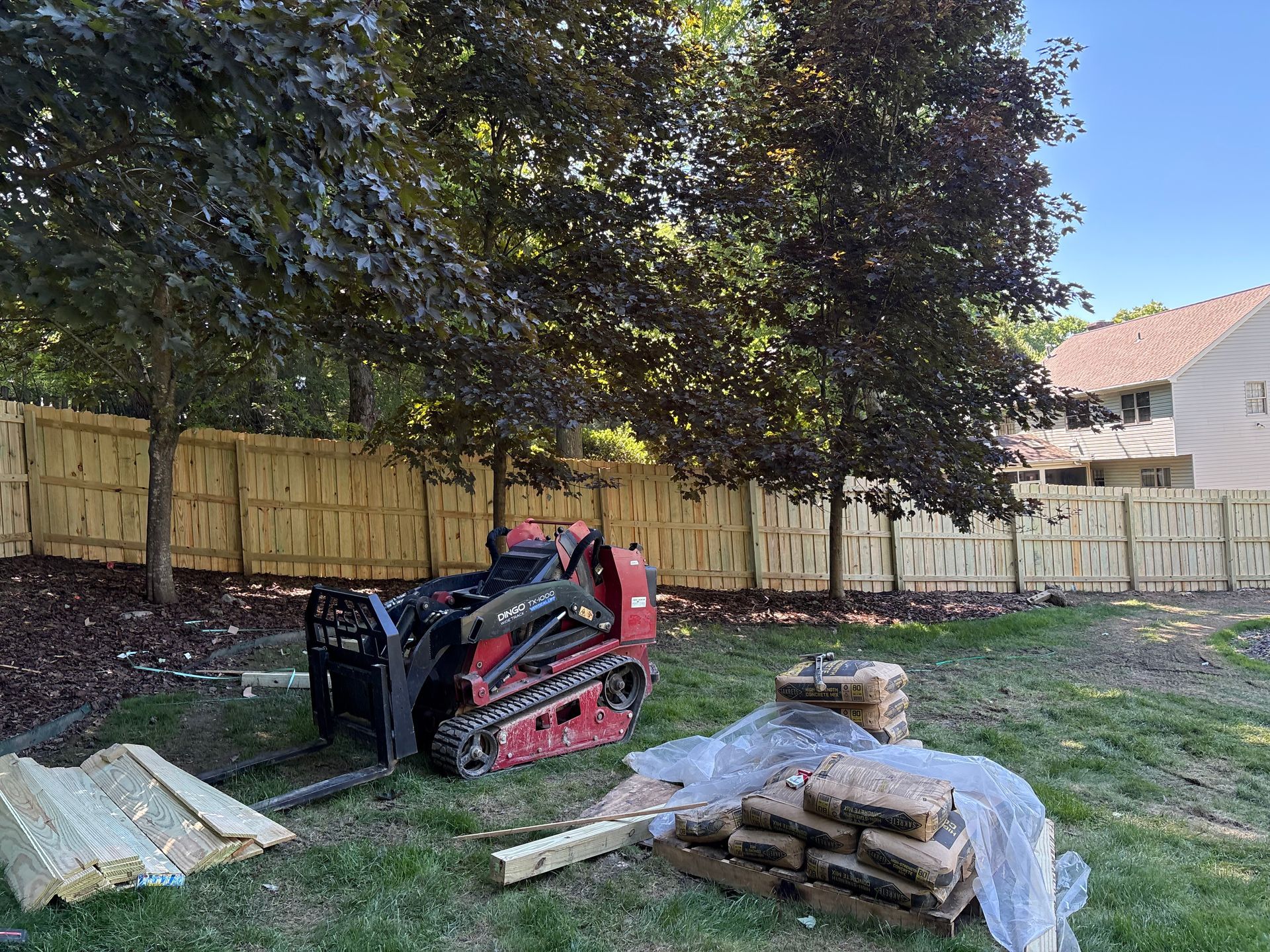 Compact track loader in a backyard beside lumber piles and a wooden fence under trees.