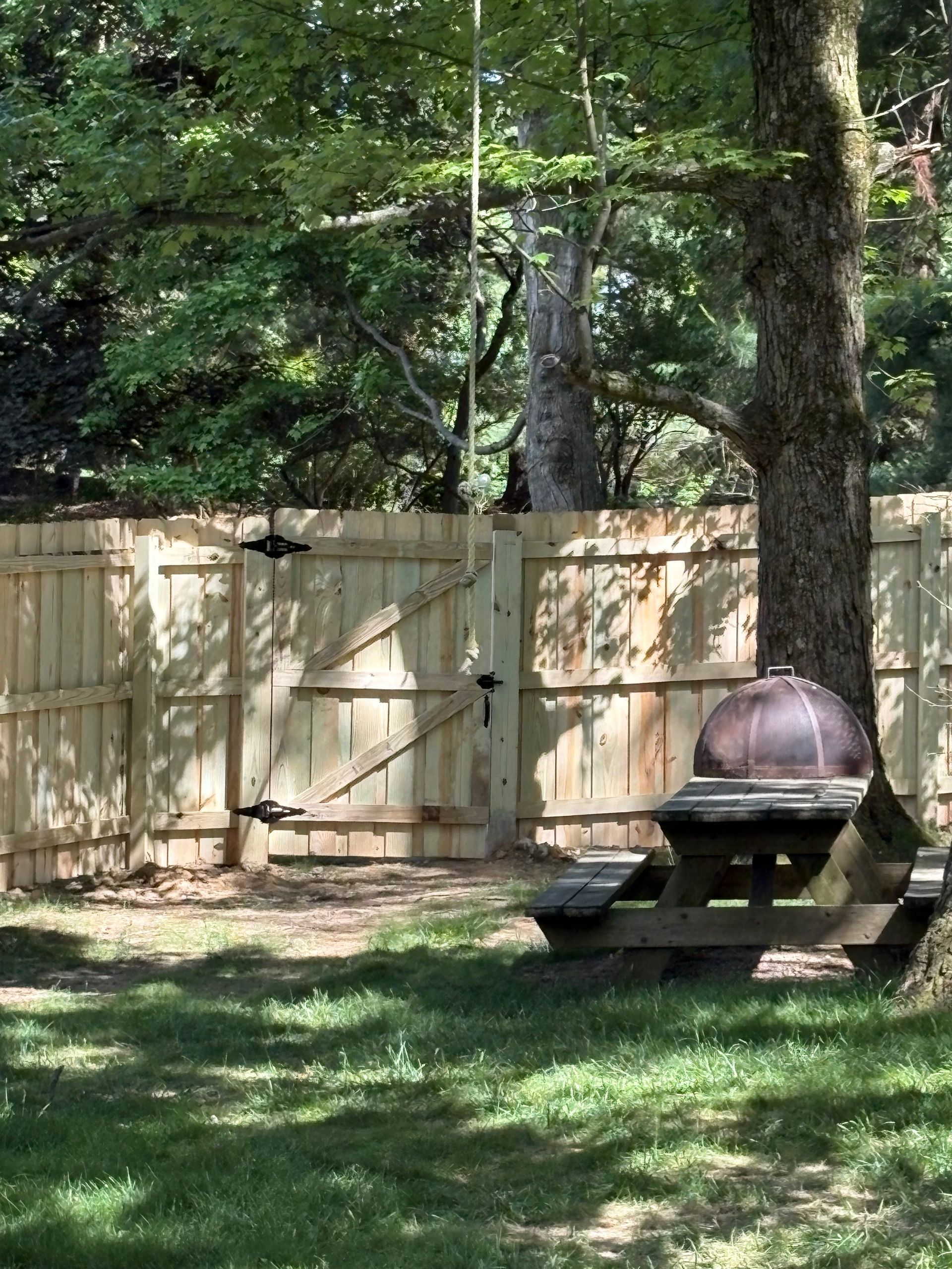 Backyard with wooden fence, trees, grass, and a picnic table beside a covered grill.