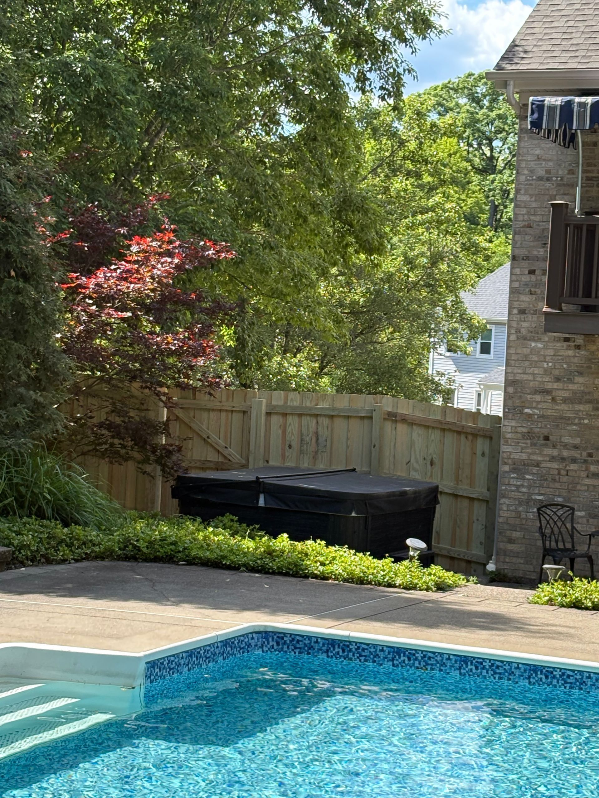 Backyard pool with blue water, stone patio, wooden fence, trees, and a hot tub beside a house