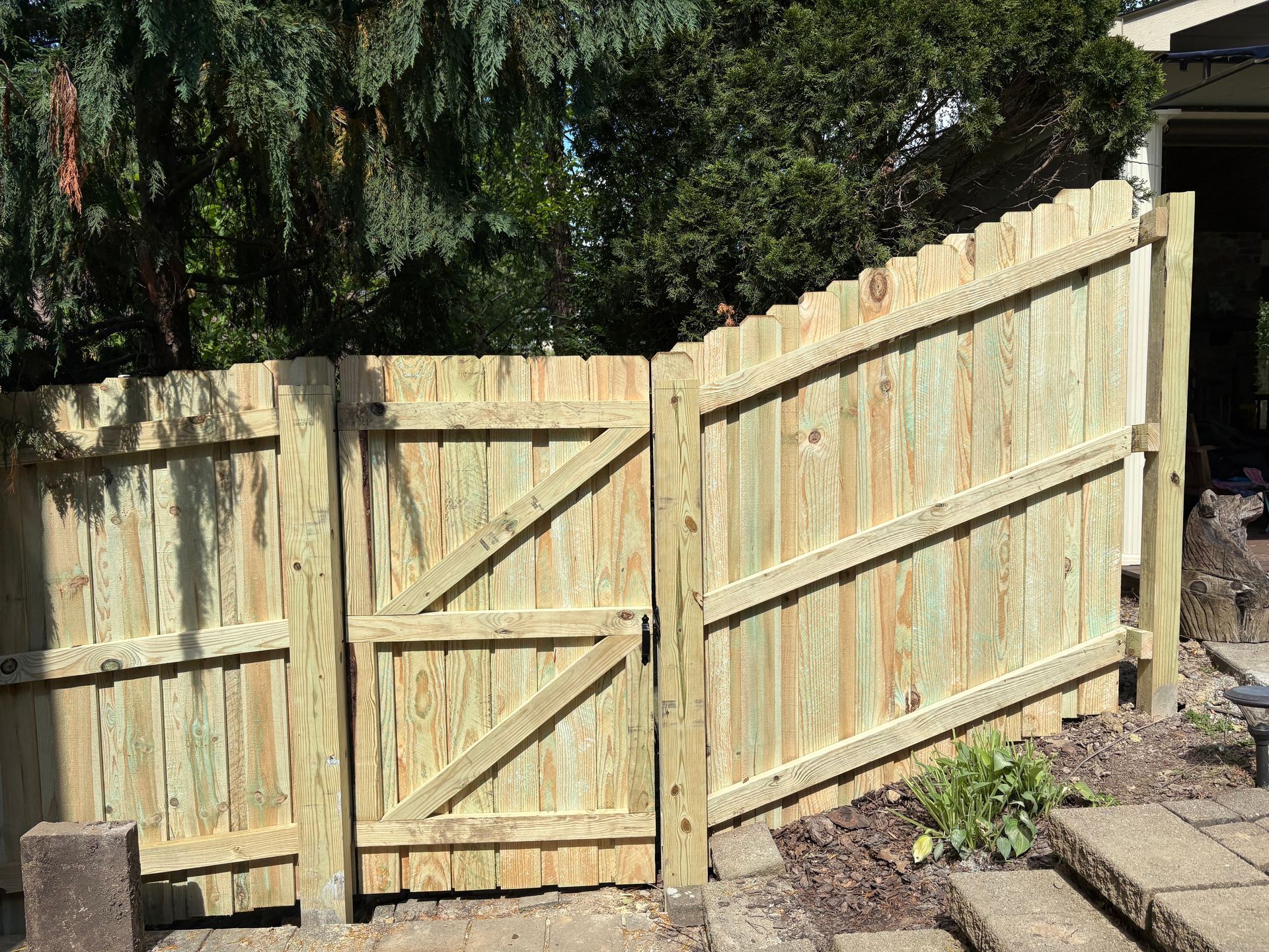 Weathered wooden backyard fence with a gate and slanted panels beside paving stones and trees.