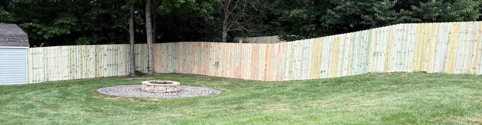 Backyard with a wooden fence, green lawn, and a small circular fire pit at center