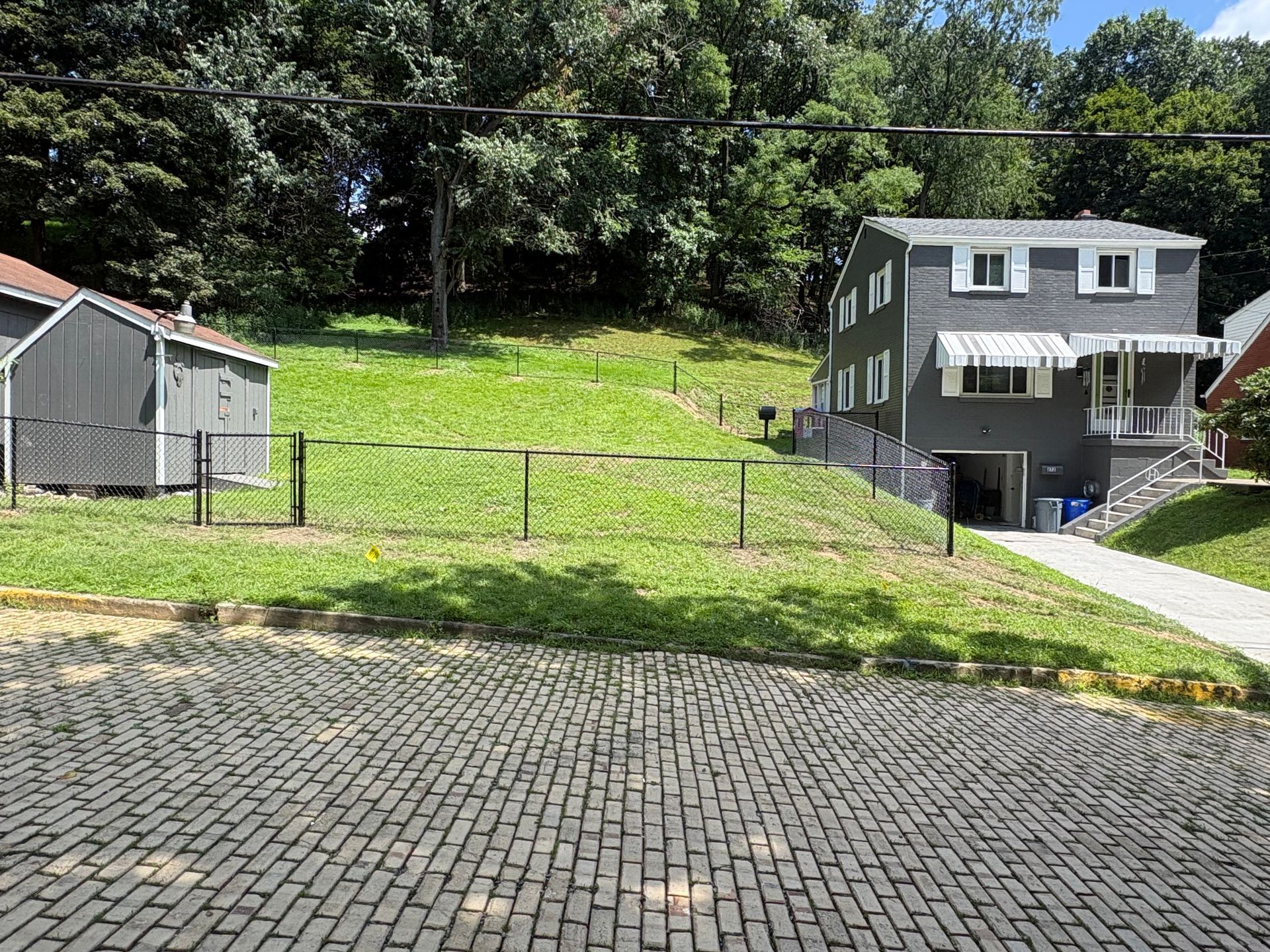 Residential hill with two houses, a grassy slope, and a paved brick driveway under trees.