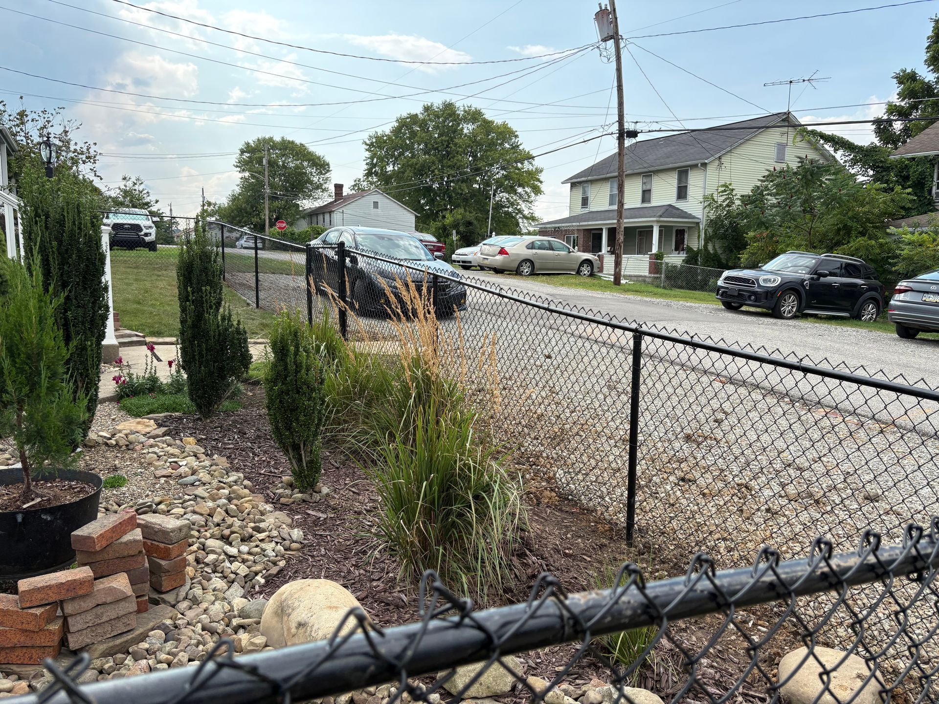 Residential street seen from a fenced front yard with parked cars and a cloudy sky