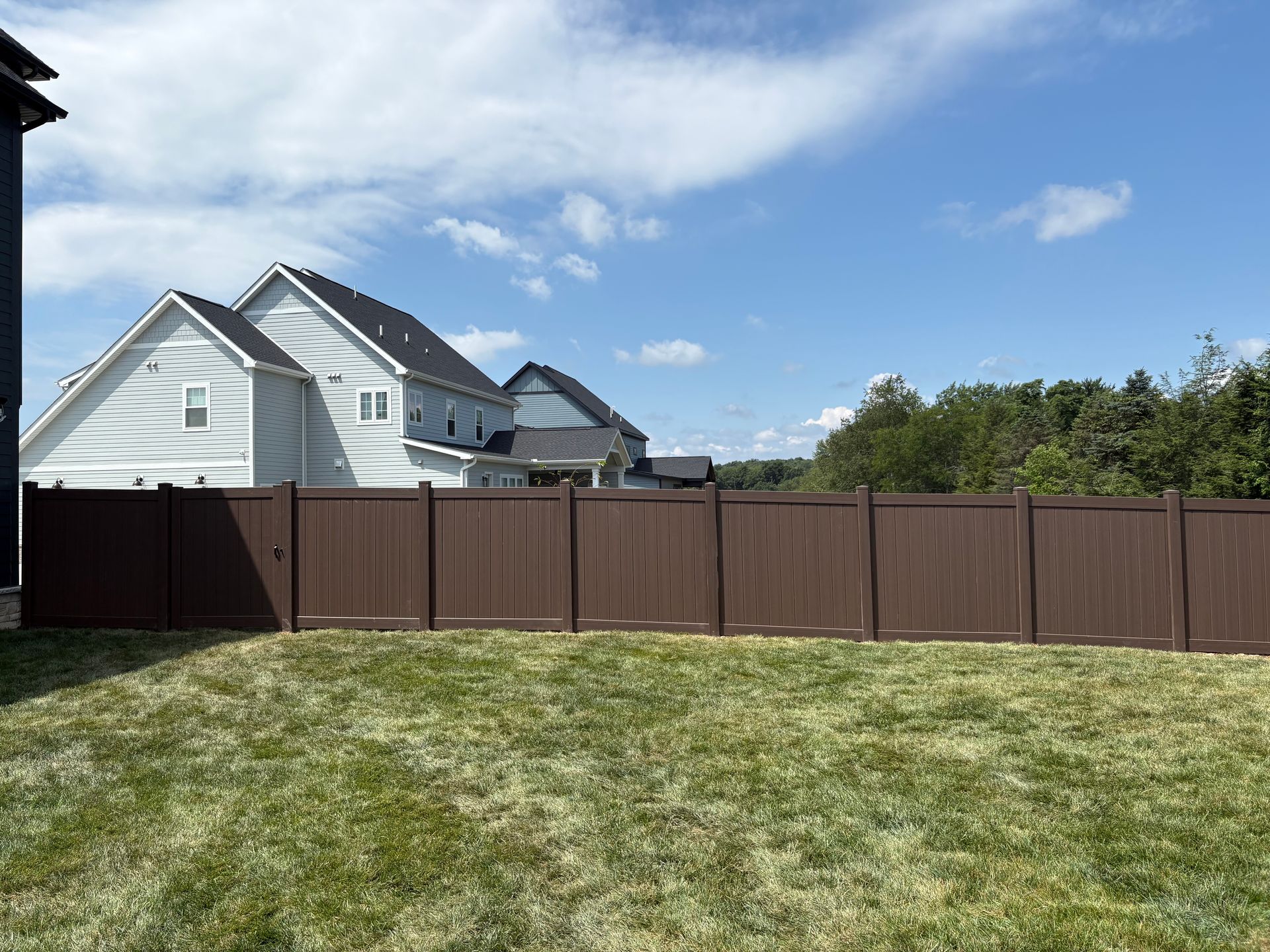 Backyard with a brown wooden fence, green grass, white house, and blue sky with clouds.