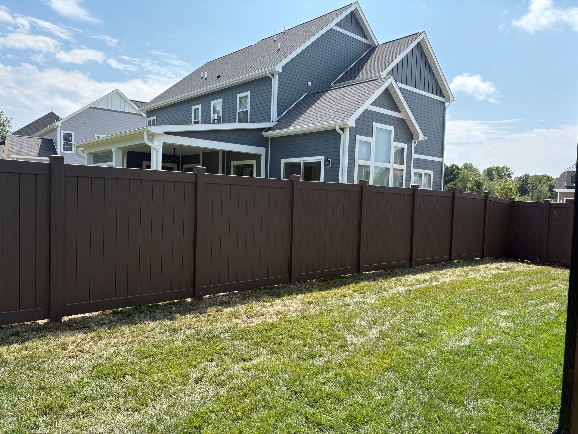 Backyard with dark brown fence, green grass, and a gray house under a blue sky