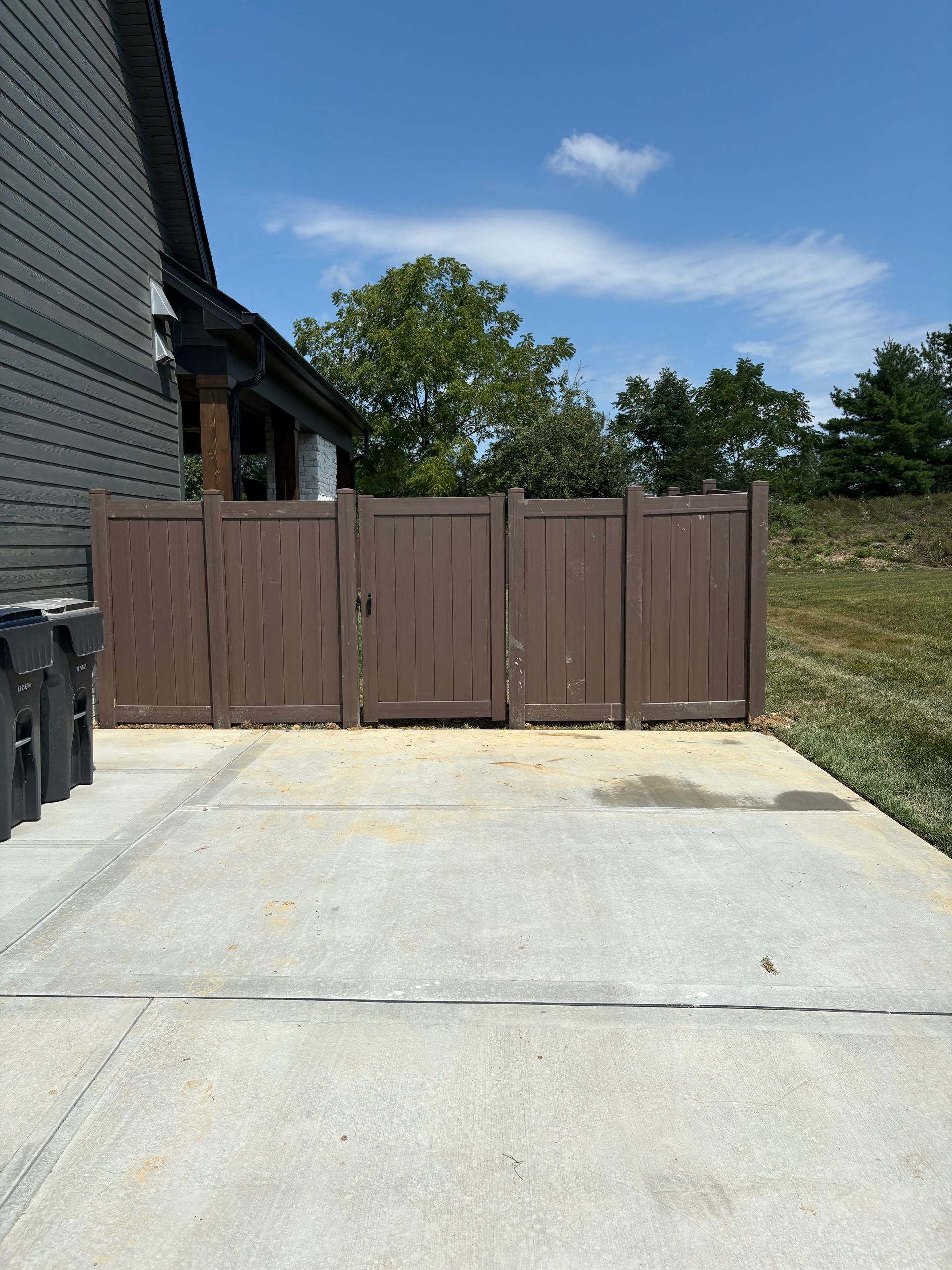 Driveway leading to a brown wooden fence beside a gray house under a blue sky.
