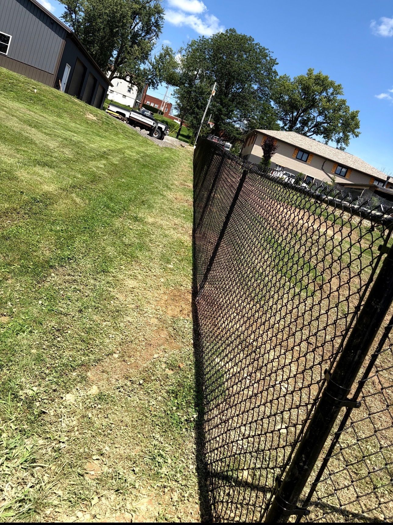 Tilted view of a black chain-link fence beside a grassy slope and apartment buildings under a blue sky