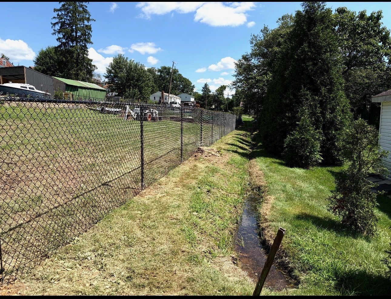Chain-link fence beside a grassy drainage ditch on a sunny suburban street with houses and trees