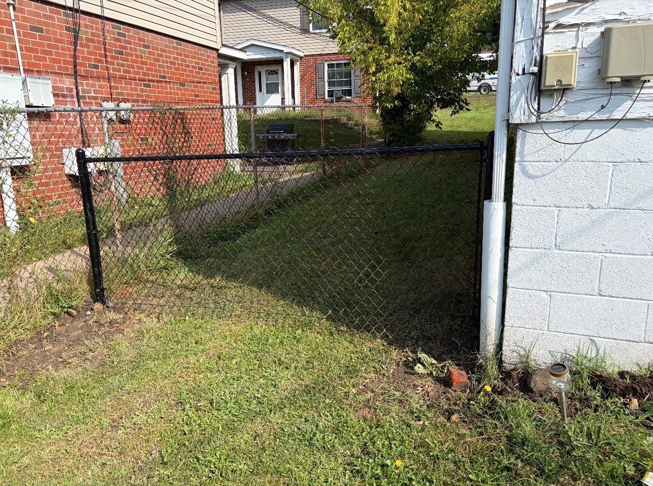 Narrow grassy alley beside a brick house and white utility building, with a chain-link fence and backyard view