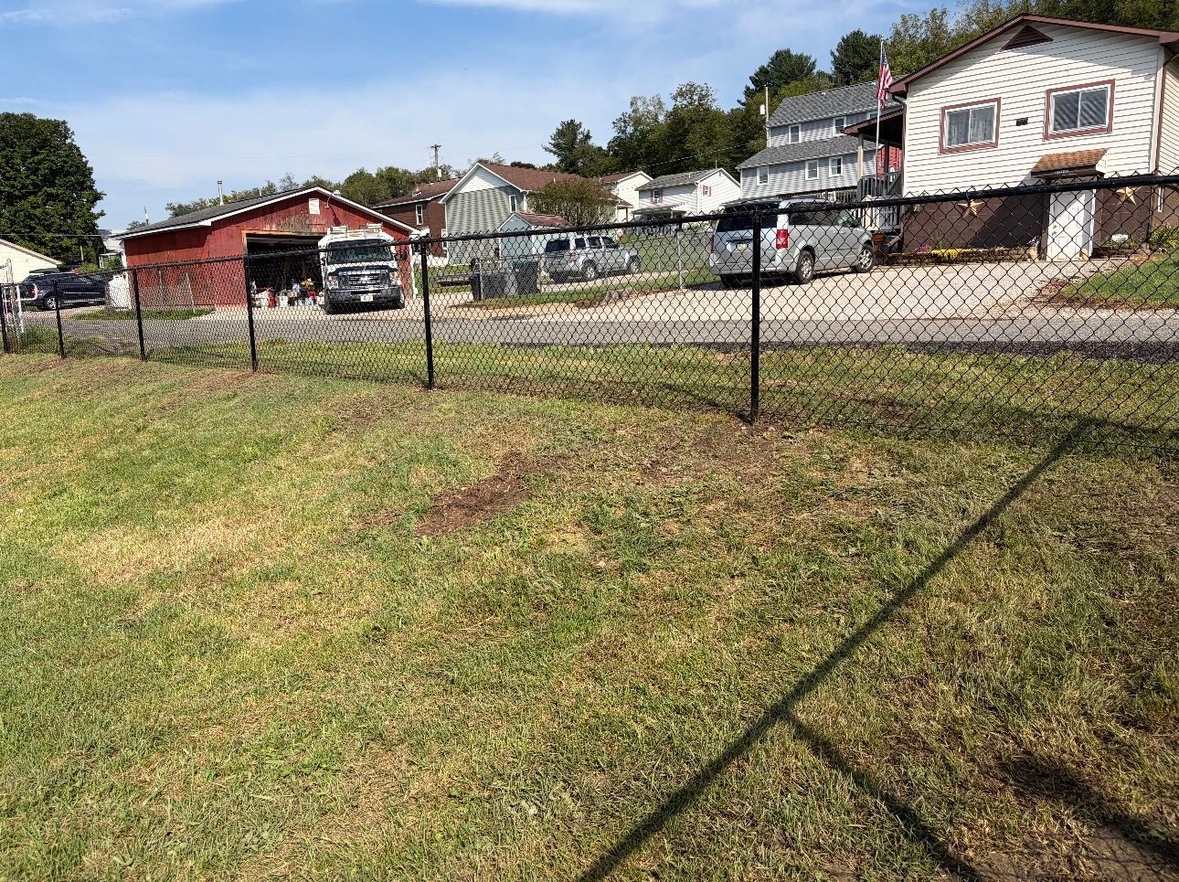 Grassy yard with a chain-link fence, houses, parked vehicles, and a red shed on a sunny day
