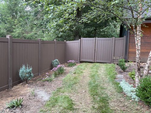 Backyard garden beside a brown wooden fence, with a gate, grass, and shrubs.