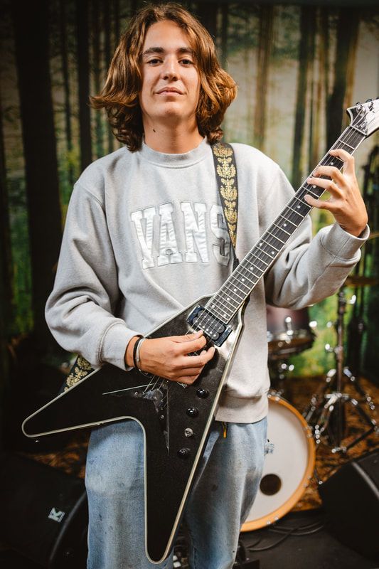 A young man is holding a guitar in front of a drum set.