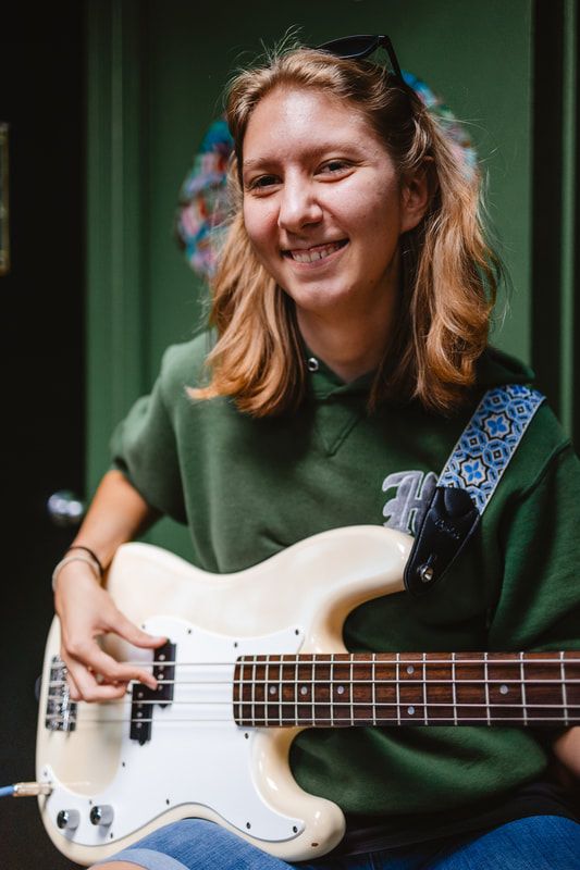 A woman in a green sweatshirt is holding a white bass guitar.